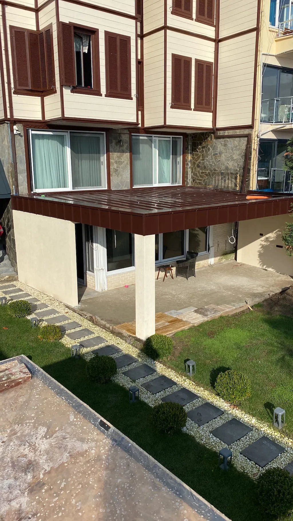 Ahşap görünümlü cephe kaplaması yenilenen villanın arka bahçesi ve metal saçaklı yeni teras gölgeliği